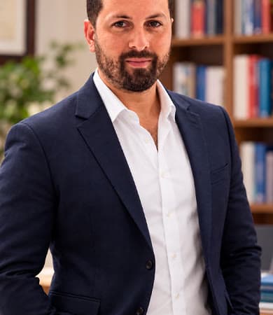 Retrato de Bruno Tavares, de terno azul-marinho, em um escritório com estante de livros